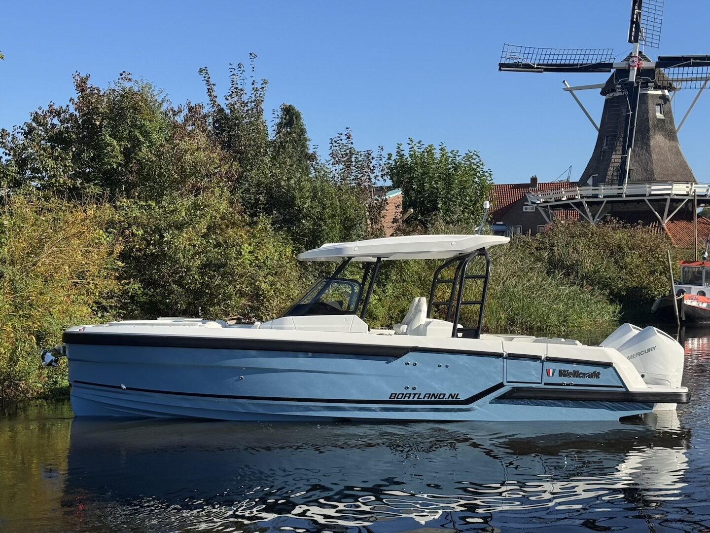 Lichtblauwe Wellcraft 28 T-Top met Mercury-buitenboordmotor afgemeerd langs een begroeide oever, met een windmolen op de achtergrond